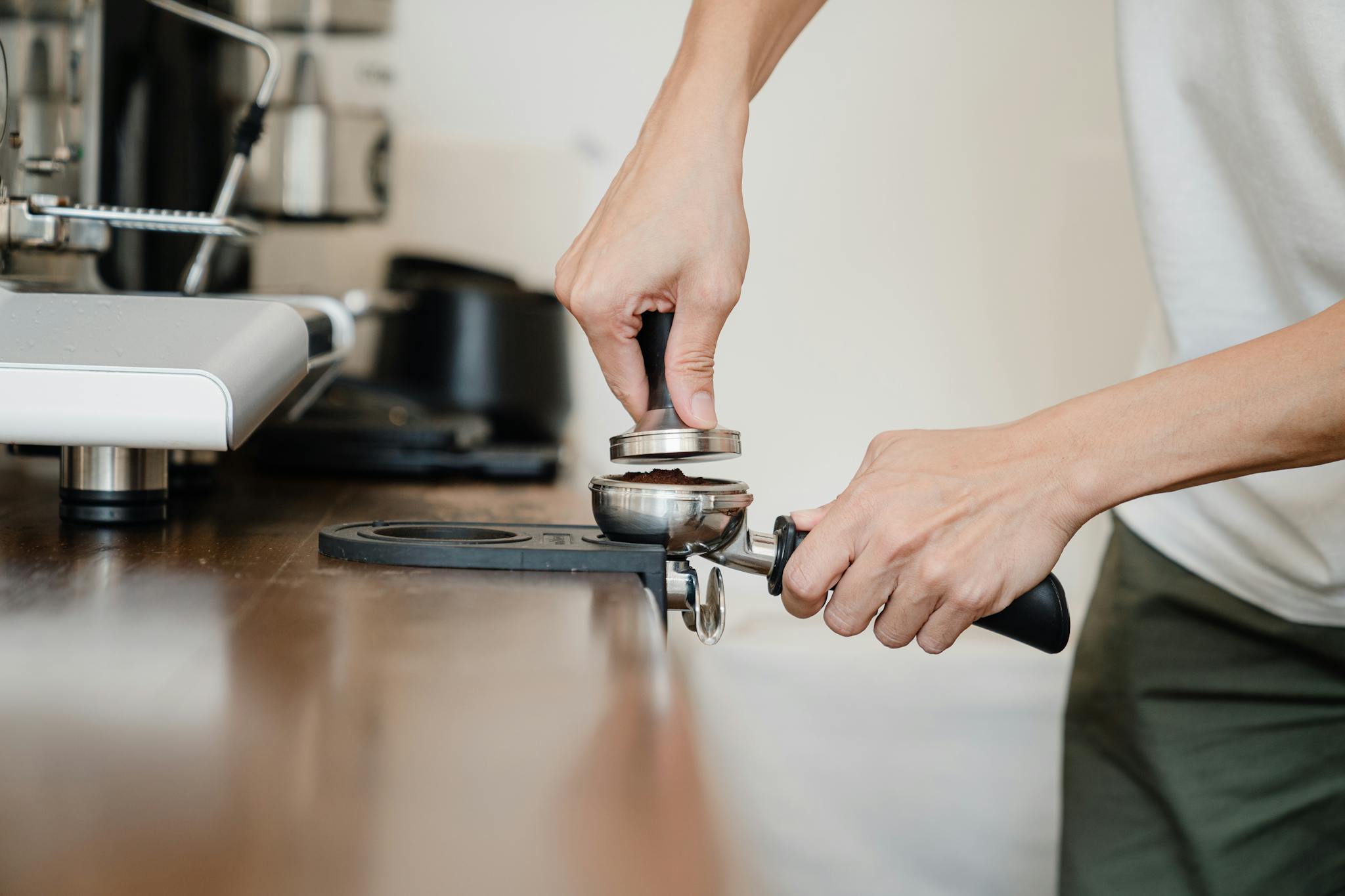 Crop anonymous male barista in casual clothes pressing ground coffee into metal portafilter by using tamper while working in contemporary kitchen